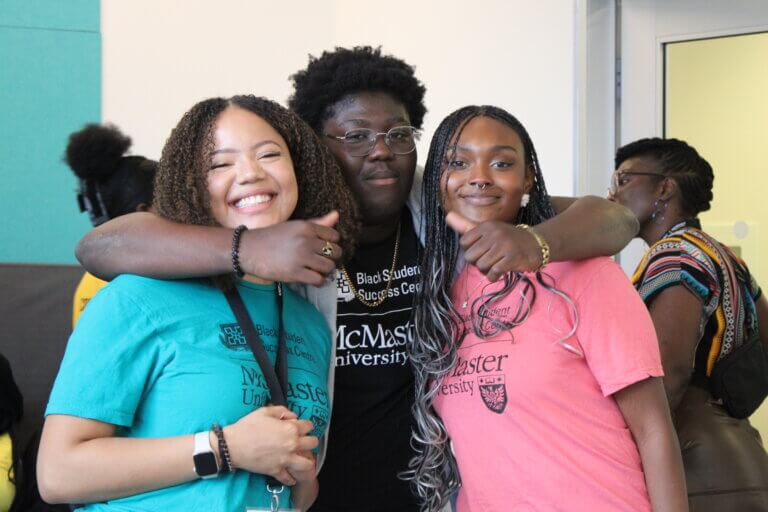 Group of three smiling friends in colorful shirts, embracing at an indoor university event. The atmosphere is joyful and welcoming.