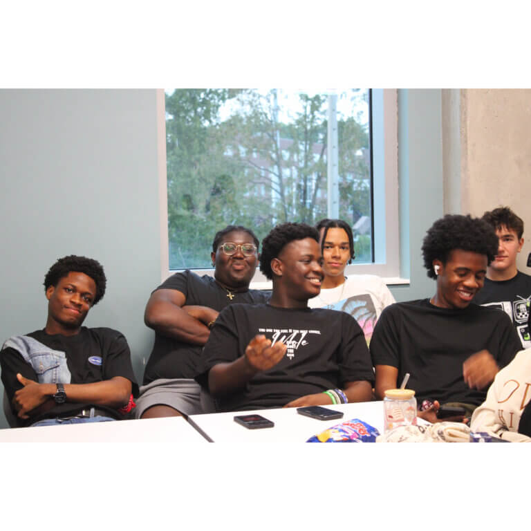 A group of seven young people sit around a table, laughing and smiling. The mood is joyful and relaxed. A window reveals trees outside.