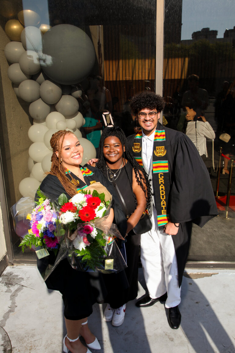 Three people celebrate a graduation. One holds flowers, another wears a cap and gown with a kente stole. They smile in front of festive balloons.