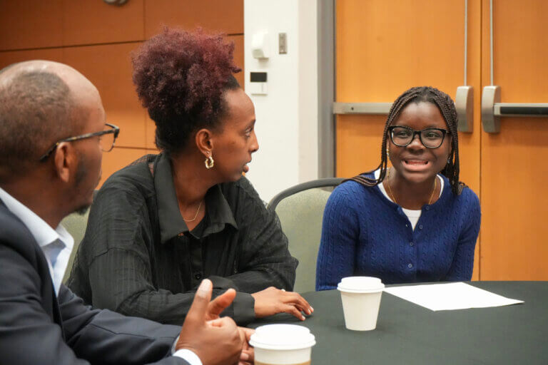 Three people sit at a table with coffee cups, engaged in a lively conversation. The setting is professional and the mood is positive and engaged.