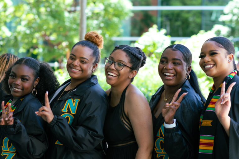 Five women smile and pose outdoors, flashing peace signs. They wear sorority jackets and stoles, celebrating an achievement with joy and camaraderie.
