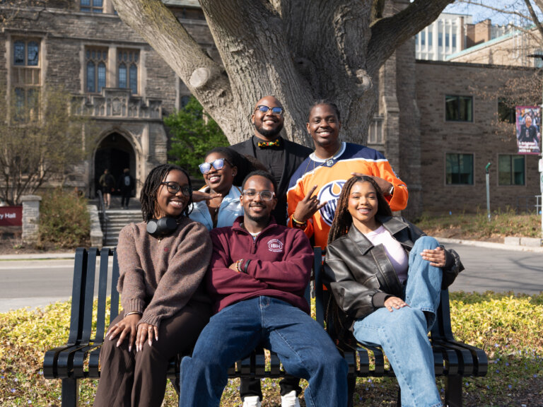 A diverse group of six people sits and stands cheerfully on a bench under a large tree. They smile warmly, capturing a friendly and upbeat mood.