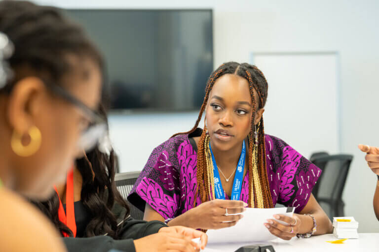 A woman in a purple patterned top sits at a table, holding papers and attentively listening to a colleague in a meeting room. Her expression is focused.