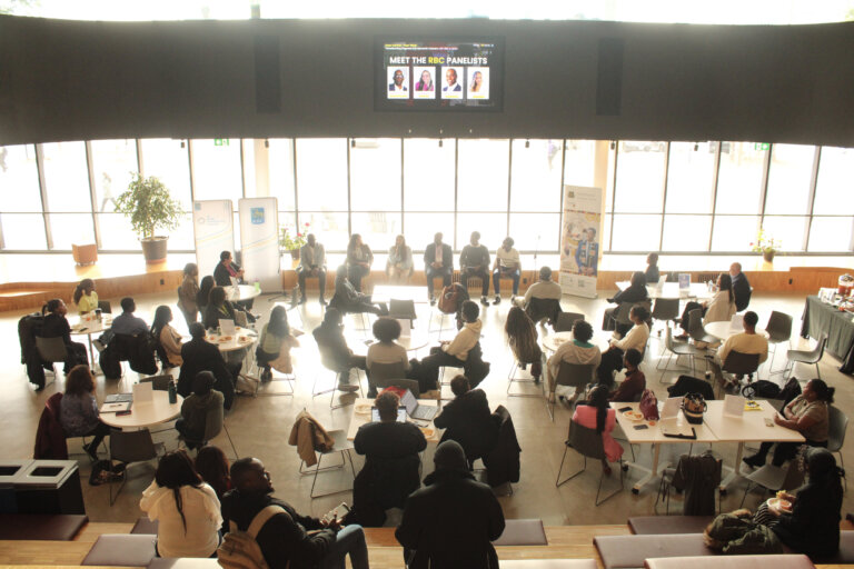 A panel discussion with eight speakers sits in front of a large audience in a modern, well-lit room. Attendees seated at round tables create an engaging atmosphere.