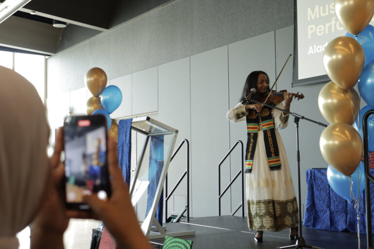 A woman in traditional attire performs on a violin on stage decorated with gold and blue balloons. An audience member captures the moment on a phone.