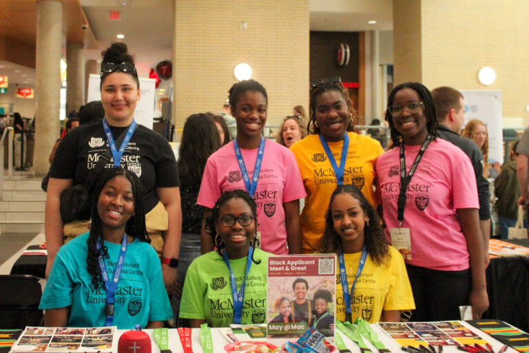 A group of seven smiling students wearing colorful McMaster University t-shirts and lanyards stand behind a table at a university fair. The table displays brochures and a sign for a 