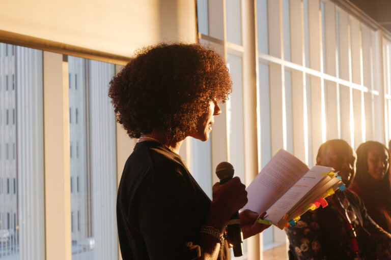 A person with curly hair speaks into a microphone, holding a book with sticky notes. They are in a sunlit room, with large windows in the background.