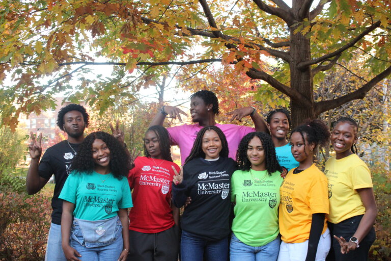 A diverse group of smiling students in colorful McMaster University tees pose under autumn trees with vibrant foliage, exuding a joyful, inclusive vibe.