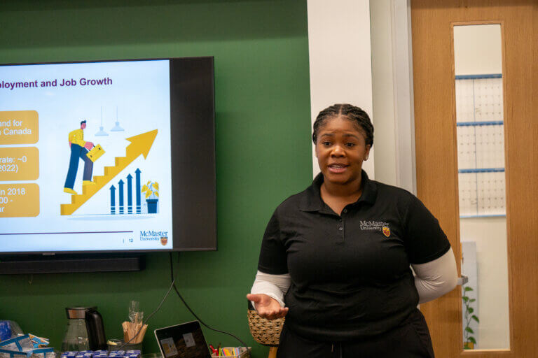 A woman presents in a classroom, standing next to a screen displaying a job growth chart. She looks engaged, conveying confidence and focus.