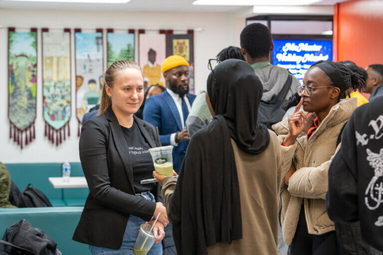 A diverse group of people in a lively setting, engaged in conversation while holding drinks. Background features colorful wall hangings. Casual, social atmosphere.