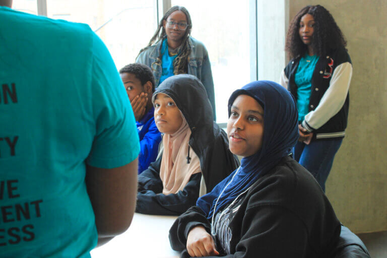 A group of engaged students sits attentively at a table, listening to a presenter. Two are in hijabs, with an attentive and curious atmosphere.