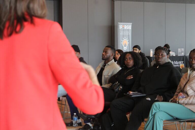 A person in a red jacket speaks to a focused, seated audience in a seminar room. A banner in the background reads 