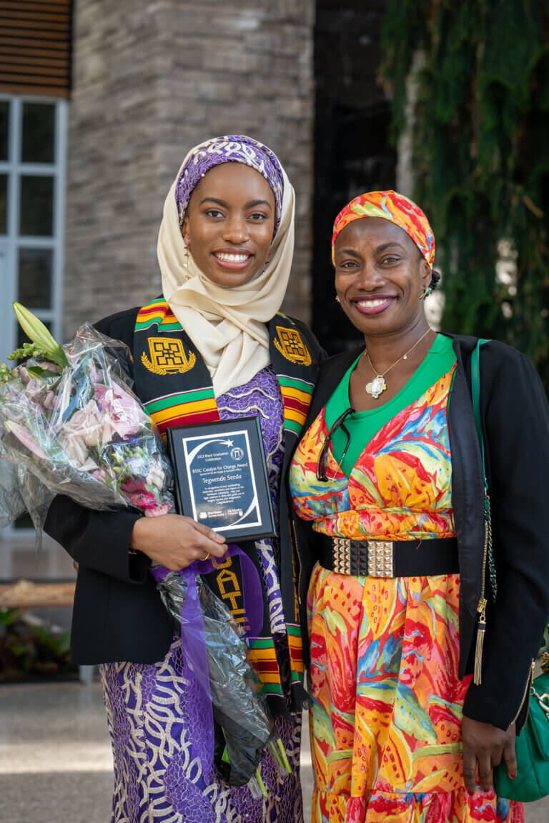 Two women smiling warmly, one in a colorful dress and headscarf, the other in a hijab and graduation sash, holds flowers and a plaque. Graduation celebration.