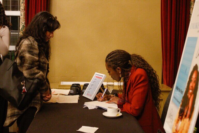 A woman in a red jacket signs a book for a person with a large bag at a table, suggesting a book signing event. A poster and coffee cups nearby.