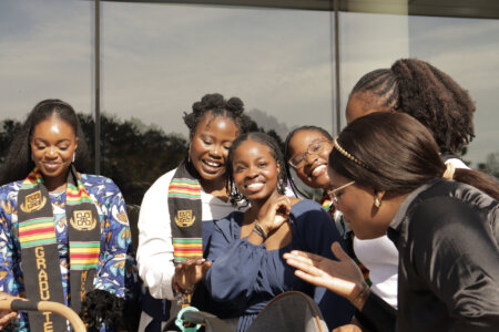 A group of seven young women celebrate outdoors. They are smiling, wearing graduation sashes, and appear joyful and celebratory.