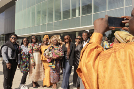 A group of smiling people posing for a photo outside a modern building. One person is holding flowers, while another wearing orange captures the moment.