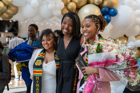 Three women smiling at a graduation celebration, wearing colorful stoles. One holds a bouquet. They're surrounded by gold and white balloons.