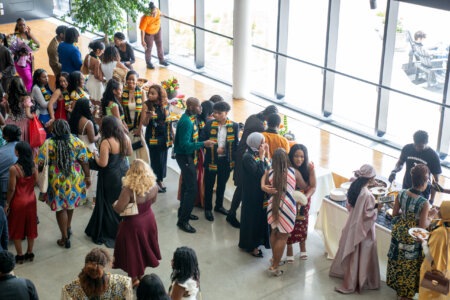 A lively graduation reception with diverse attendees. Graduates in colorful stoles and formal attire mingle near a buffet by large windows, evoking celebration.
