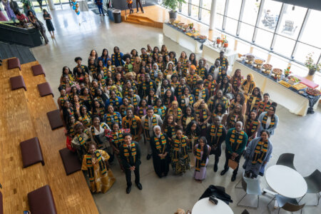 A large group of people wearing traditional stoles are gathered in a bright, spacious hall with wooden seating and tables set up with food, conveying a joyful and celebratory atmosphere.