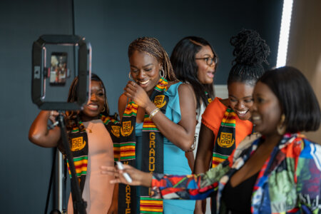 A group of women wearing colorful stoles and smiling joyfully in front of a smartphone on a tripod. They're celebrating, exuding happiness and pride.