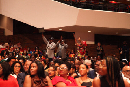 A lively audience in an auditorium, with some people standing and clapping, wearing colorful attire. The mood is celebratory and engaged.